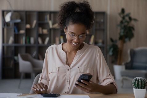 Cobranças recorrentes podem acelerar seu negócio e suas vendas. Mulher utilizando a calculadora e sorrindo ao olhar para o smartphone.